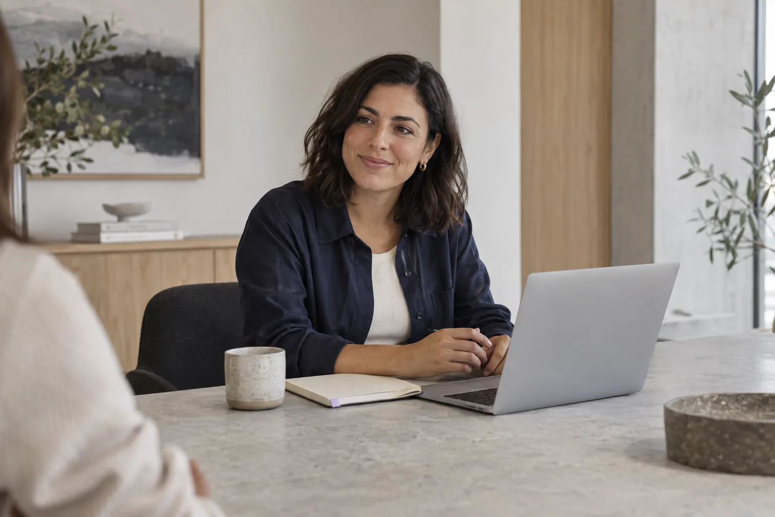 A woman in conversation beside an open laptop in a light-filled studio.
